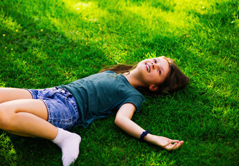Happy laughing fun casual kid girl lying on the grass and looking up on the sky on nature summer sunny brigh background. Closeup - 792229347