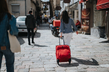 A young woman in a striped shirt pulls a red suitcase along a vibrant cobblestone street in a...