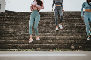 Three women in athletic attire actively exercising by running up concrete steps in a city setting,...