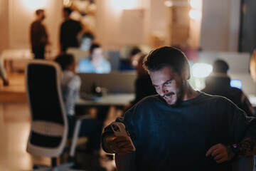 Happy young male with facial hair smiling while using his smart phone, with colleagues working in...
