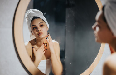 Fototapeta na wymiar Fresh and clean young woman with a towel wrapped on her head standing in front of a bathroom mirror, reflecting daily skincare routine.