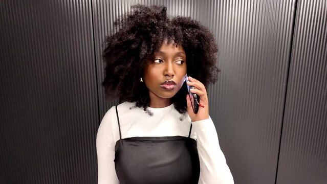 A contemporary young black woman is engaged with her smartphone, texting and making calls in what appears to be the confines of an urban elevator. Her focused expression and stylish appearance capture