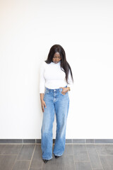 A young Black woman strikes a relaxed pose against a minimalist white wall. Her long, sleek hair complements the timeless elegance of a fitted white turtleneck, paired perfectly with chic wide-leg