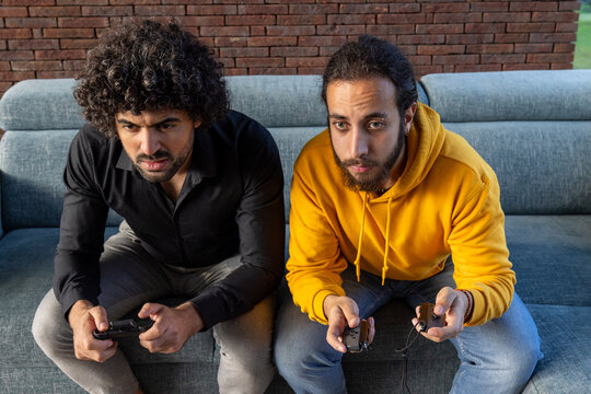 Two engaged adult males are seated on a grey couch, holding game controllers with intense concentration. They are indoors, with a red brick wall in the background, implying a casual, home environment