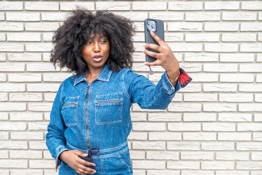 A confident young Black woman with natural afro hair takes a selfie with her smartphone. She's wearing a stylish denim jumpsuit against a white brick wall backdrop. Her expression is playful as she