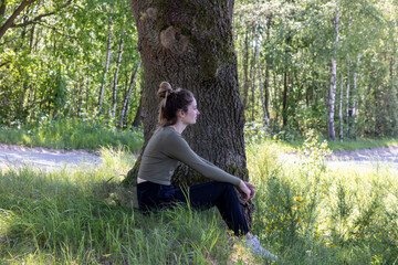 This image eloquently captures a young woman in a contemplative state, sitting against an old tree's sturdy trunk. Her profile is set against a backdrop of dense birch trees and lush undergrowth, with