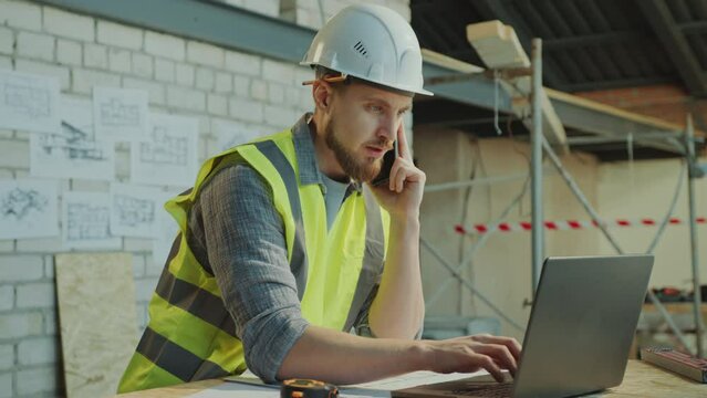 Male engineer wearing hardhat and safety vest typing on laptop, looking at floor plans and having conversation on mobile phone at construction site