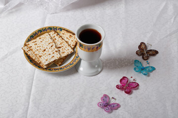 Matzah, unleavened bread on a plate and wine cup both ornately decorated. Four butterflies add a touch of whimsy. The setting for Passover is on a subtly patterned white tablecloth. Judaism, Religious