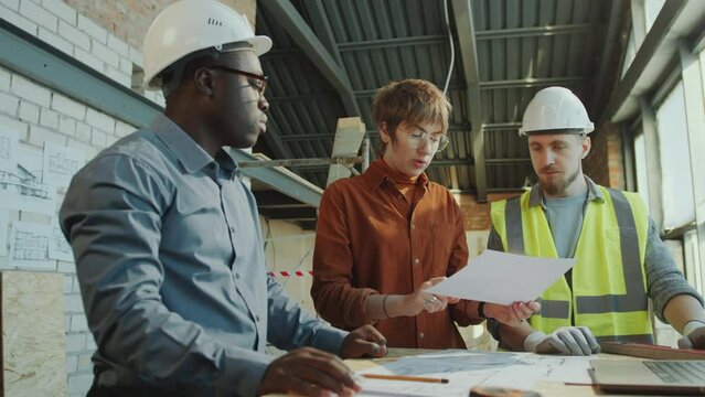 African American architect and Caucasian engineer wearing hardhat discussing building plan on paper with female client visiting construction site