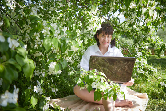 Girl Working on Laptop in Blossoming Garden. Middle aged woman using computer in white apple blossoms. Female freelancer, blogger working outdoors