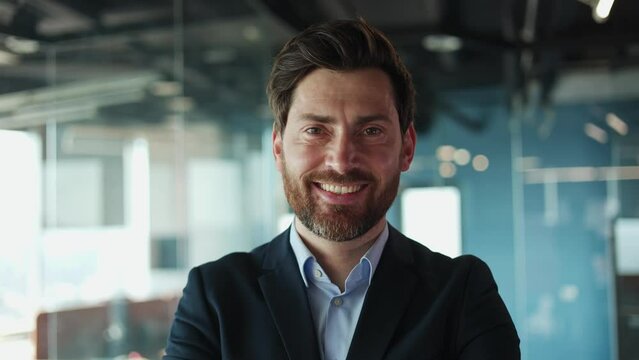 Close up of positive caucasian male smiling widely while standing against translucent expanse of office wall. Attractive businessman wearing dark formal suit showing resolve in the workplace.