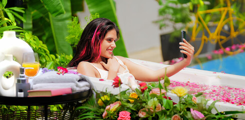 Indian Asian smiling gen z young adult girl sitting rose petal tub take selfie use mobile cam hold...