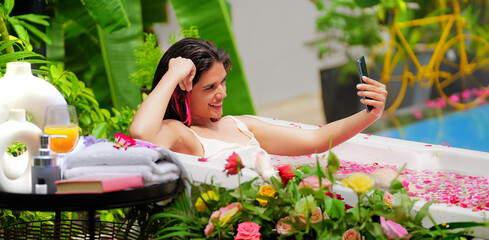 Indian Asian smiling gen z young adult girl sitting rose petal tub take selfie use mobile cam hold phone home. Happy cute wet woman pose look cam make video call enjoy fun joy hotel villa resort trip - obrazy, fototapety, plakaty