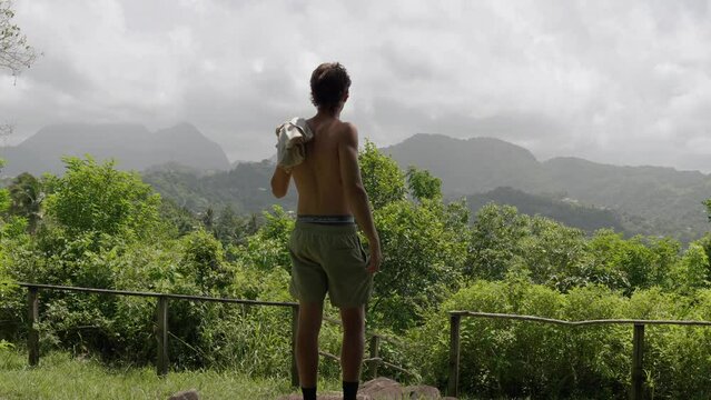 Shirtless man taking shirt off shoulder while standing in front of a tropical mountainous jungle in the Caribbean on Saint Lucia island.