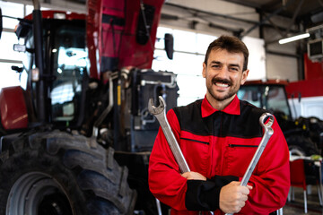 Portrait of smiling mechanic standing by the tractor inside workshop maintaining agricultural...