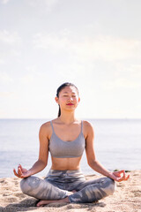 Fototapeta na wymiar young asian woman doing meditation at beach sitting with legs crossed, concept of mental relaxation and healthy lifestyle, copy space for text