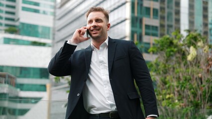 Successful businessman standing at rooftop while talking with marketing team about investment by using phone. Caucasian investor enjoy relaxing by looking at modern skyscraper view. Lifestyle. Urbane