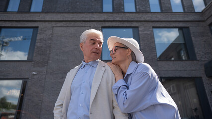 A woman in a hat and glasses hugs an elderly man in a white jacket from behind. Romantic relationships of mature people. 