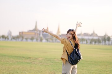 A Traveler Asian woman in 30s, bathed in golden glow of a Bangkok sunset, laughs with carefree joy, her arms outstretched as if to embrace freedom of the moment. Backpack strapped on. - 792477936