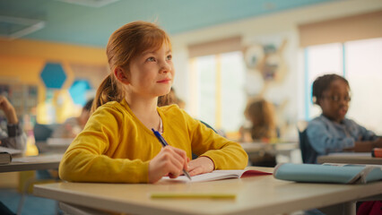 Portrait of a Cute Smiling Girl with Bright Ginger Hair Sitting Behind a Desk in Class in Elementary School. Young Pupil is Focused on a Lecture, Listening to a Teacher with Other Kids