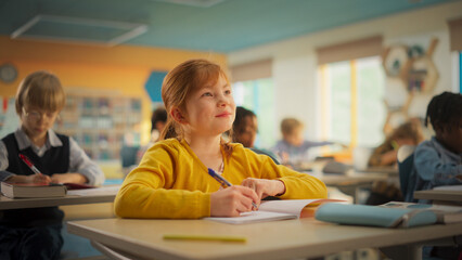 Smart Little Redhead Girl in Bright Clothes Sitting Behind a Desk in Primary School. Young...