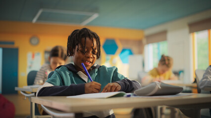 Portrait of a Cute Little African Boy with Stylish Hair Sitting Behind a Desk in Class in Elementary School. Young Pupil is Focused on a Lecture, Listening to a Teacher with Other Kids