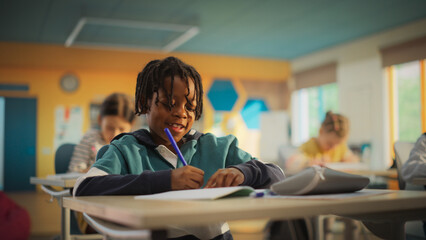 Portrait of a Cute Little African Boy with Stylish Hair Sitting Behind a Desk in Class in Elementary School. Young Pupil is Focused on a Lecture, Listening to a Teacher with Other Kids - Powered by Adobe