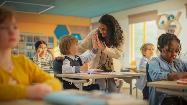 Enthusiastic Female Teacher Walking Between Rows, Checking Test Assignment Results, High Fiving with an Adorable Smart Boy. Class of Multiethnic Children Listening to a Lecture and Writing Down Notes