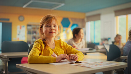 Portrait of a Cute Smiling Redhead Girl Sitting Behind a Desk in Class in Elementary School. Young Pupil is Looking at Camera, Smiling. Kids Educated in Modern Primary School