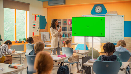 Primary School Children Having an Information Hour in Class: Female Teacher Educating Smart Diverse Kids in a Modern Colorful Classroom, Using Television with a Mock Up Green Screen
