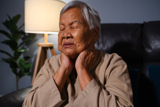 senior woman suffering from neck pain while sitting on sofa in living room at night
