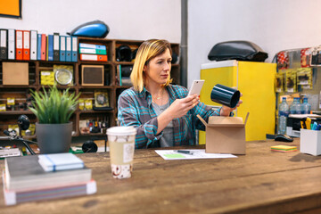 Young female entrepreneur taking photo with cell of a blue mug for client of her online store. Parcel delivery and ecommerce concept. Blonde woman reviewing new merchandise to sell in local store.