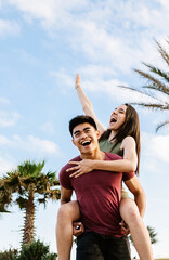 Vertical portrait of happy young multiracial couple having fun together on summer vacation. Joyful asian man piggybacking pretty woman outdoors. Love, happiness and friendship concept