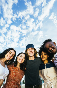 Vertical portrait of young group of happy multiracial people standing outdoors over blue sky. Cheerful multicultural friends embracing in the street. Community and unity concept. Copy space.
