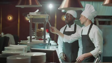 Chef walking in restaurant kitchen, attaching order ticket to metal rack and discussing it with colleague during workdayChef walking in restaurant kitchen, attaching order ticket to metal rack and dis