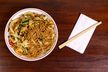 Fried noodle with chicken and vegetables in a white bowl on a wooden table. After edits.