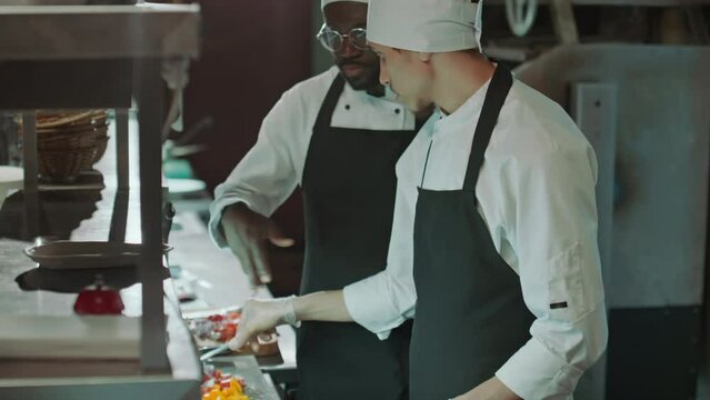Chef adding garnish to dish and teaching apprentice how to serve food in restaurant kitchenChef adding garnish to dish and teaching apprentice how to serve food in restaurant kitchenChef adding garnis
