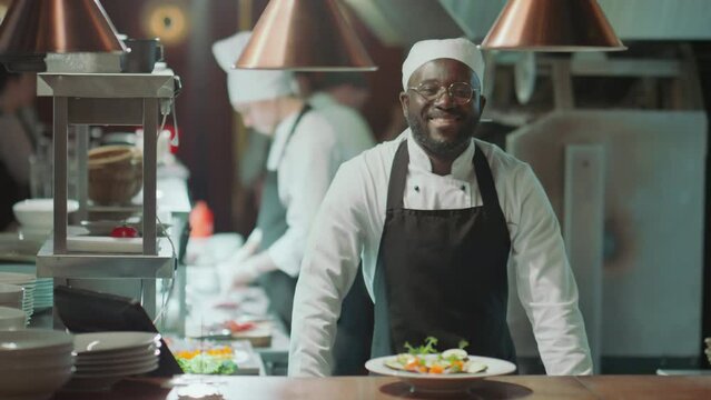 Portrait of young African American chef in apron putting plate with served dish on table and posing for camera with smile while working in restaurant kitchenPortrait of young African American chef in 