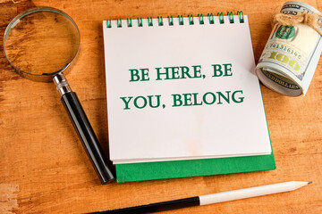 BE HERE, BE YOU, BELONG Words written on a notebook with a magnifying glass and a roll of money on a papyrus background - obrazy, fototapety, plakaty