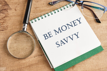 Savings concept,Text Be money savvy on a notebook with a magnifying glass and glasses on a vintage background - obrazy, fototapety, plakaty