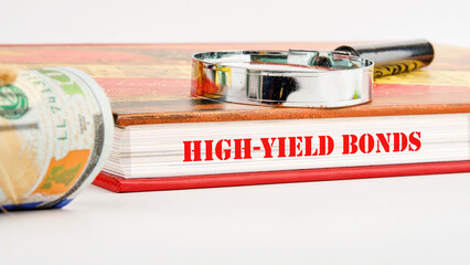 Inscription High-Yield Bonds on the pages of the book next to a roll of dollar bills and a magnifying glass on the book on a white background - obrazy, fototapety, plakaty