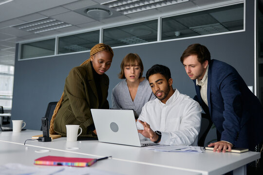 Four young adults in businesswear talking and gesturing by laptop on desk in office