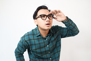 Fototapeta premium Portrait of curious asian man with hand over eyes and peering into the distance, looking away, expecting and looking for someone on the horizon. isolated studio shot