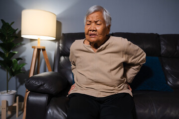 senior woman suffering from backache while sitting on sofa in living room at night
