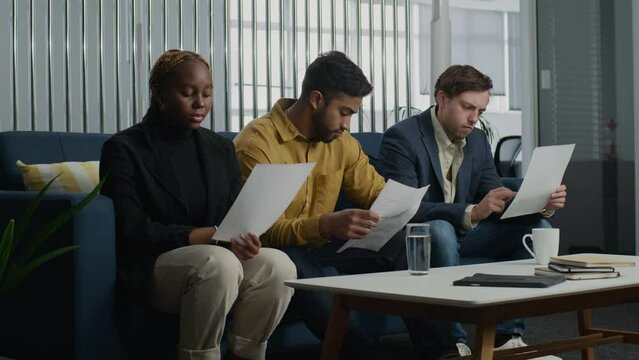 Young multiracial adults in businesswear sitting on sofa and reading paperwork in office