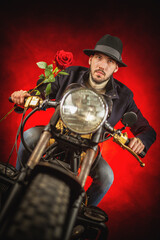 A man in classic coat  with the rose flower in hand is riding an old retro style motorcycle.