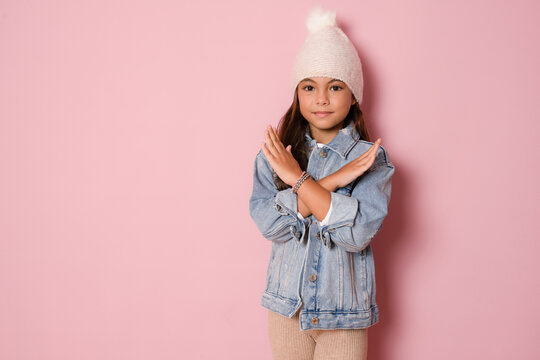 Brunette kid girl in knitted hat over pink background Has rejection angry expression on face and crossing hands doing refusal negative sign. - Powered by Adobe