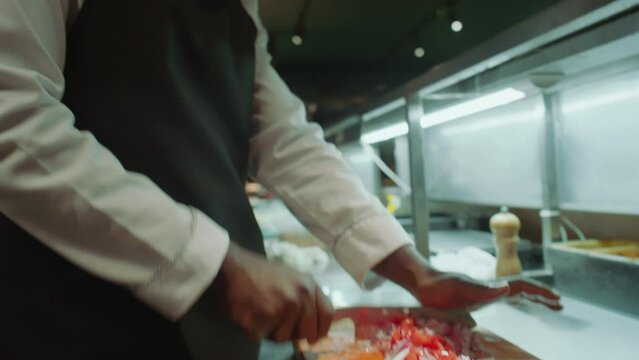 Close up shot of Black chef in apron chopping fresh tomatoes and red onion on wooden board at kitchen table while working in restaurantClose up shot of Black chef in apron chopping fresh tomatoes and 