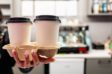 Person holding two takeaway coffee cups in a carrier, with a blurred cafe background, illustrating the convenience of takeaway coffee and on-the-go lifestyle