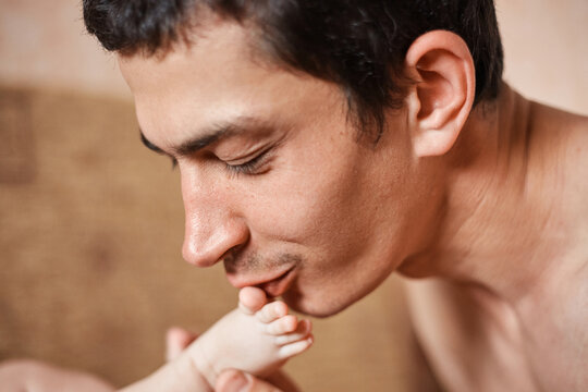 Love for the child. Closeup of brunette handsome man with newborn baby father kisses the infant kid's leg expressing love and tenderness
